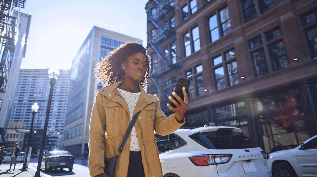 Woman in urban setting uses smartphone, sunlight reflecting on buildings.