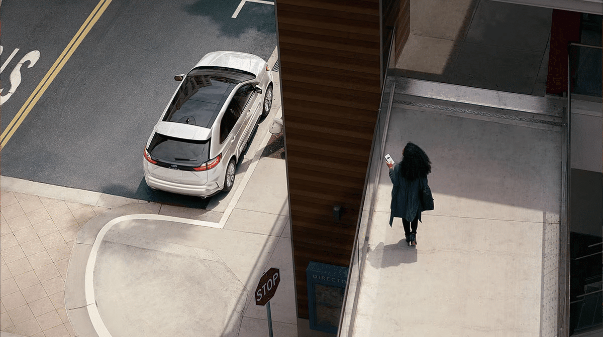Aerial view of a white car parked near a stop sign and a person with curly hair checking a phone on the sidewalk.