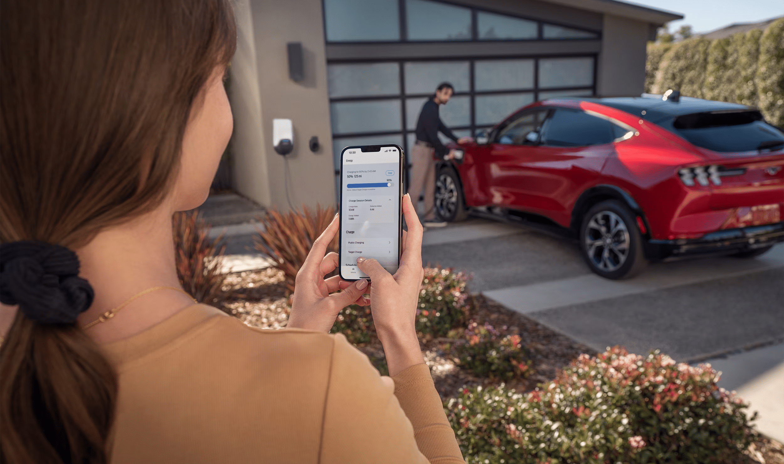 Woman using smartphone to monitor red electric car charging in driveway.