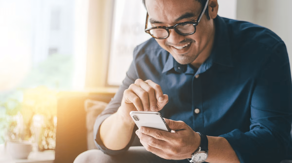 Smiling man in glasses using a smartphone in a bright, cozy room.