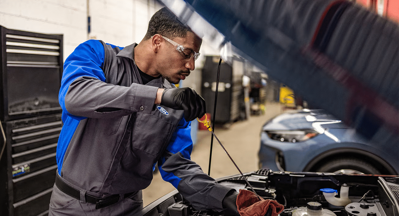 Mechanic checking engine oil with a dipstick in a car workshop, wearing safety goggles and gloves.