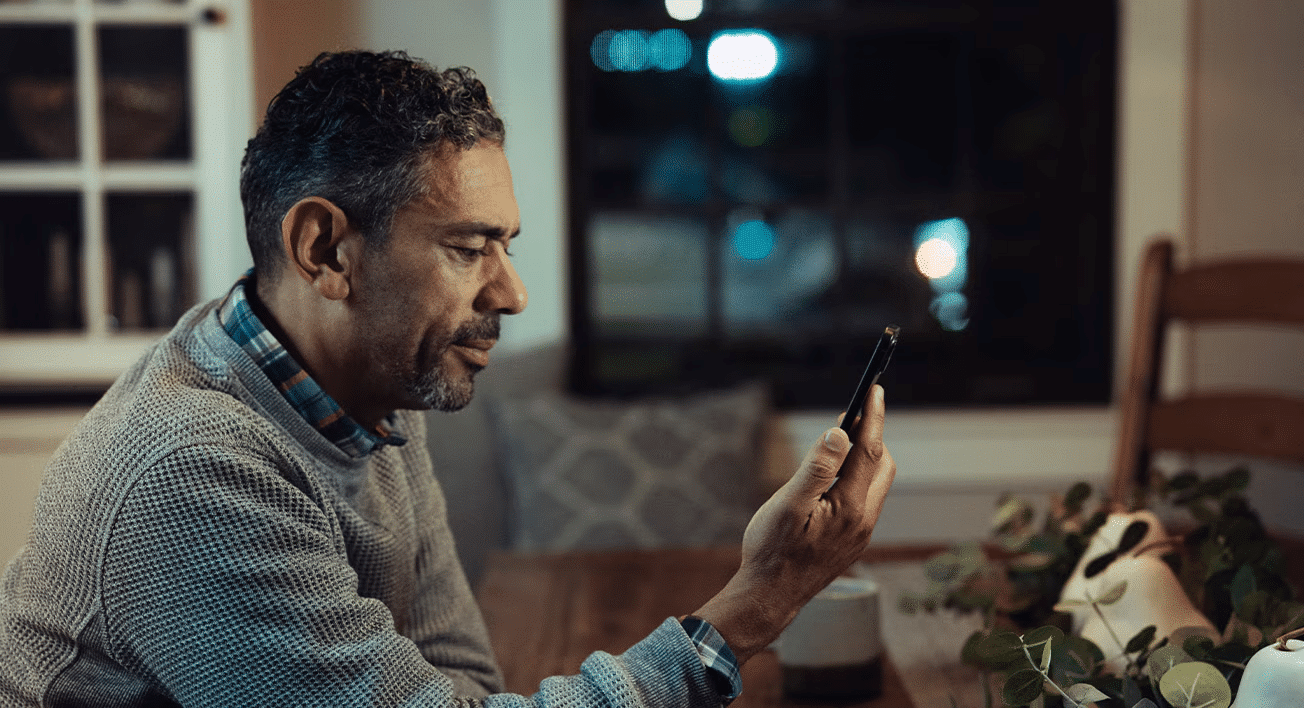 Man with gray hair intently looks at his smartphone, seated at a wooden table indoors.