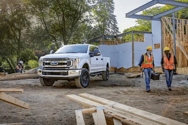 White pickup truck at a construction site with workers in safety gear carrying tools nearby.