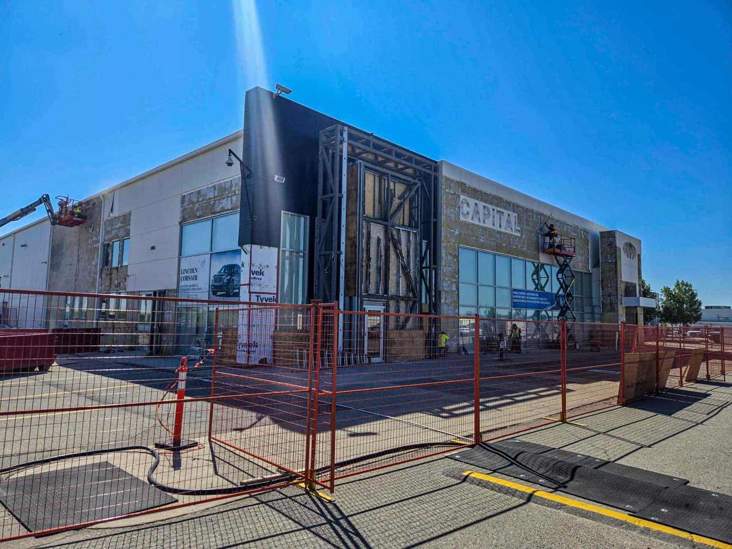 Building under renovation, surrounded by red fencing, with workers on lifts; clear blue sky above.