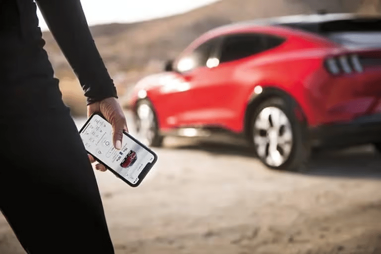 Person holding a smartphone with a red car app screen, parked red car in the background.