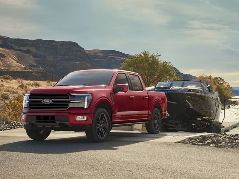 Red pickup truck towing a boat on a scenic road near rocky hills under a blue sky.