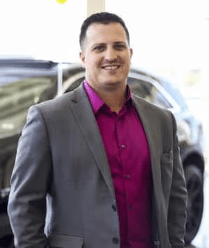 Person in a gray suit and magenta shirt smiling, standing in front of a car in a showroom.