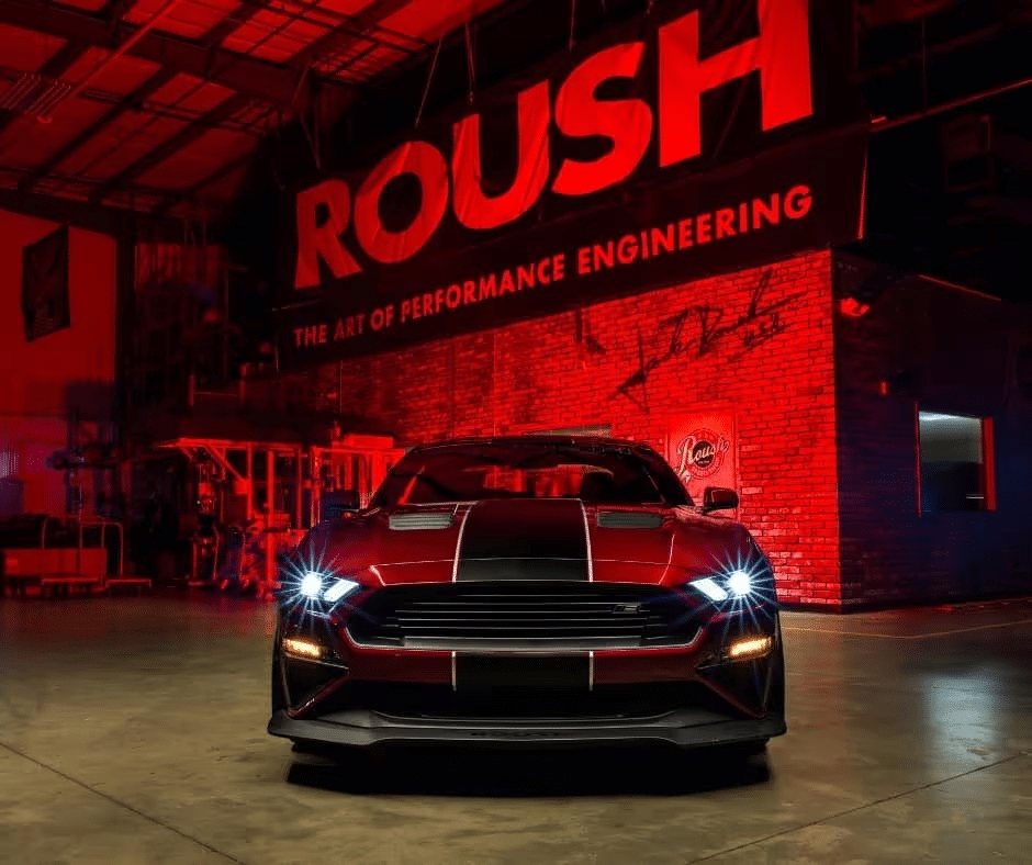Red sports car with headlights on, parked in a garage under the "ROUSH" banner.