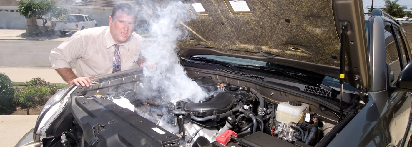 Man in a tie examining a smoking car engine on a residential street.