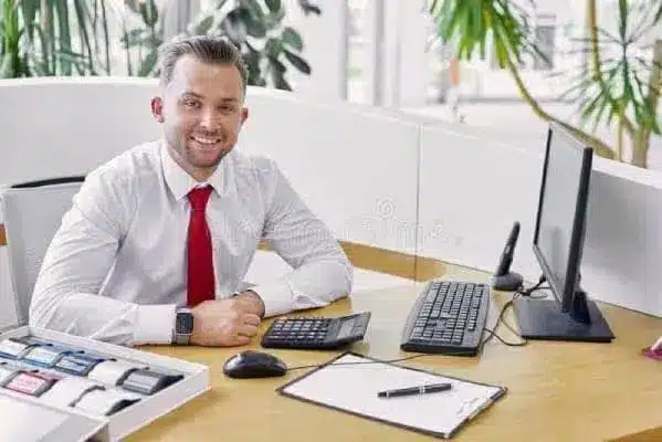 Smiling professional in a white shirt and red tie at a desk with computer, calculator, and clipboard.