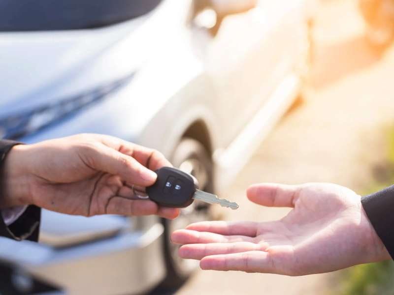 Hand giving car key to another person, with a white car in the background.