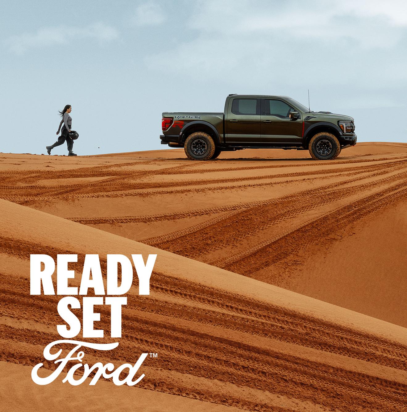 A Ford truck on a sandy desert dune with a person walking nearby. Text reads "READY SET Ford."